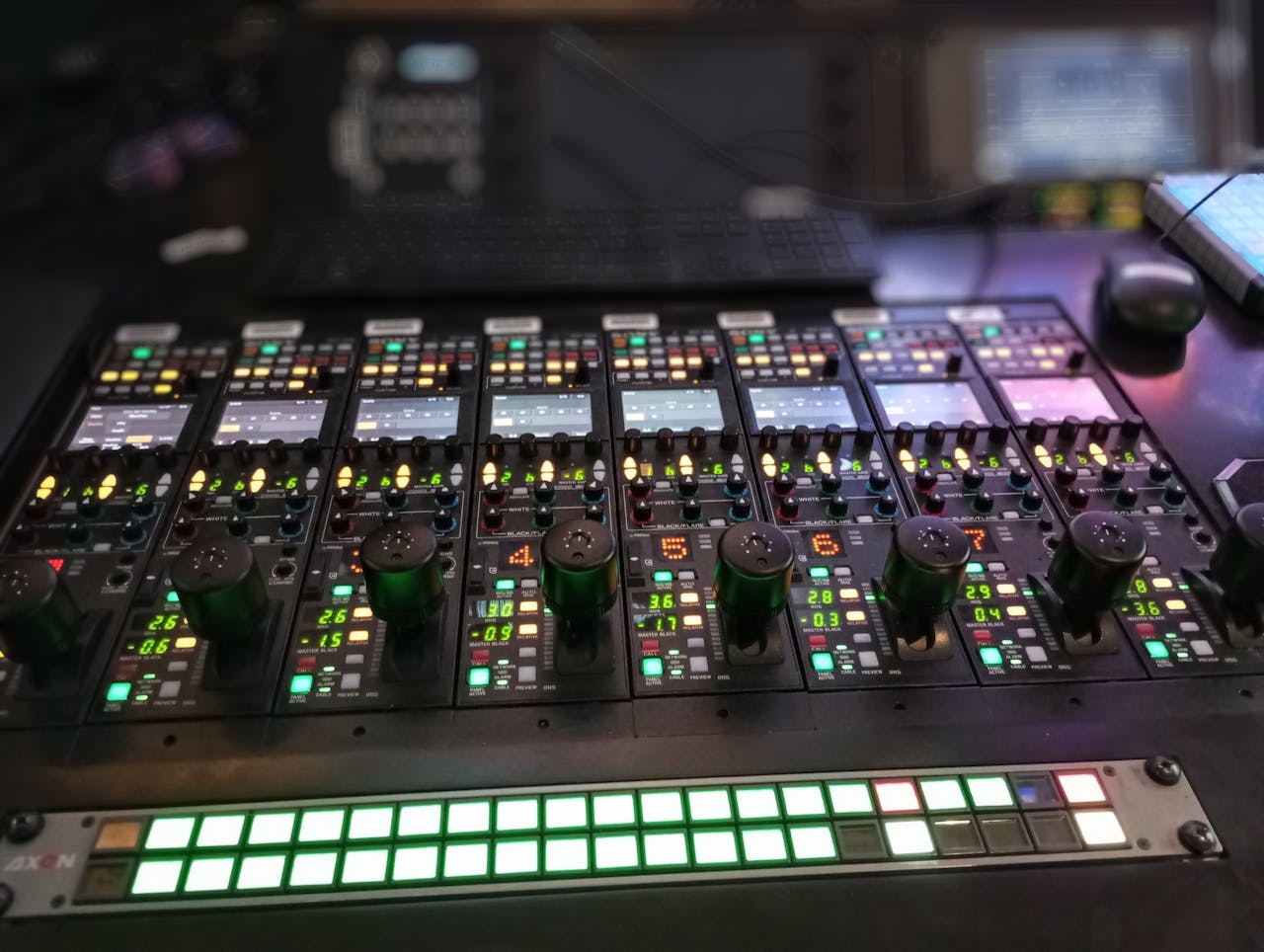 gallery-02 Close-up of an illuminated audio mixer panel in a recording studio, showcasing various controls and switches.